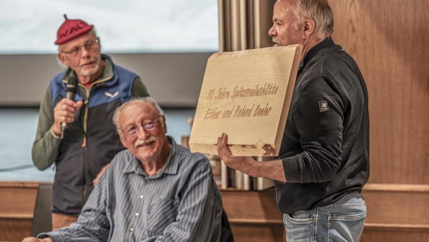 Hüttenchef Walter Brühlmann (links) und der Hüttenchef der Schräawislihütte, Robert Kohler (Mitte), überreichen dem Hüttenwartpaar der Spitzmeilenhütte, Roland und Esther Beeler, zum 10-Jahr-Jubiläum ein Präsent. Esther kümmert sich derweil um ihre Hütte. 